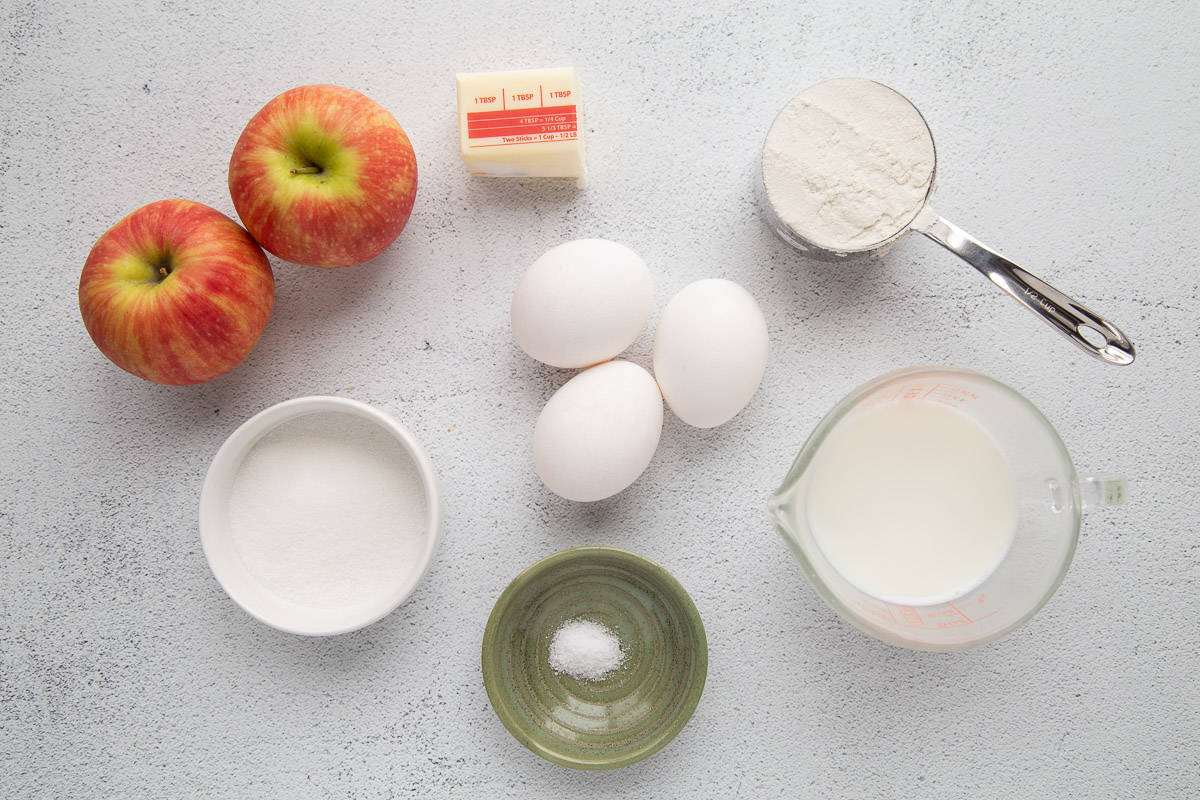 eggs, flour, milk, apples, and butter on a white table.