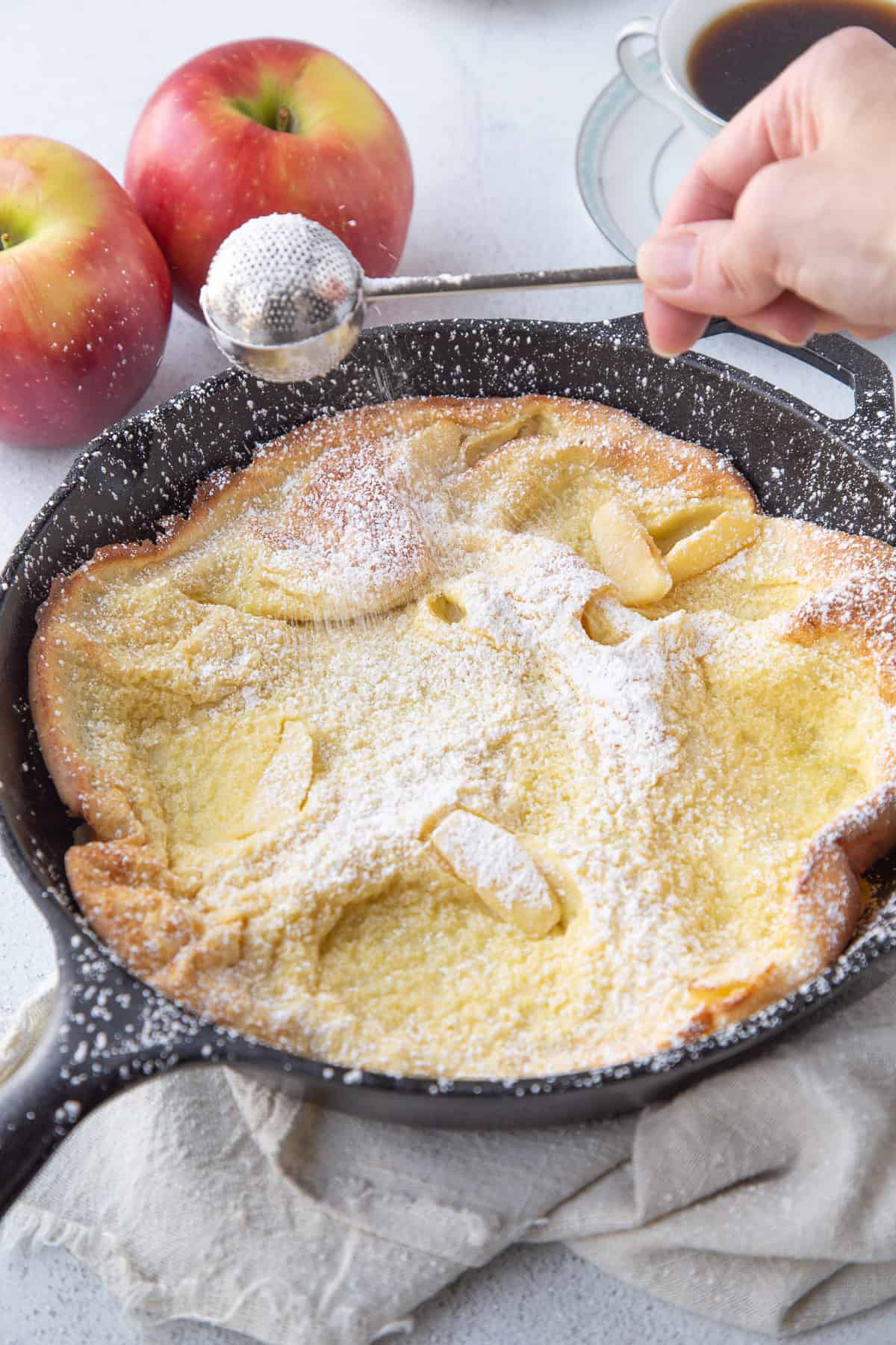 hand using a tea strainer to dust a german pancake with powdered sugar.