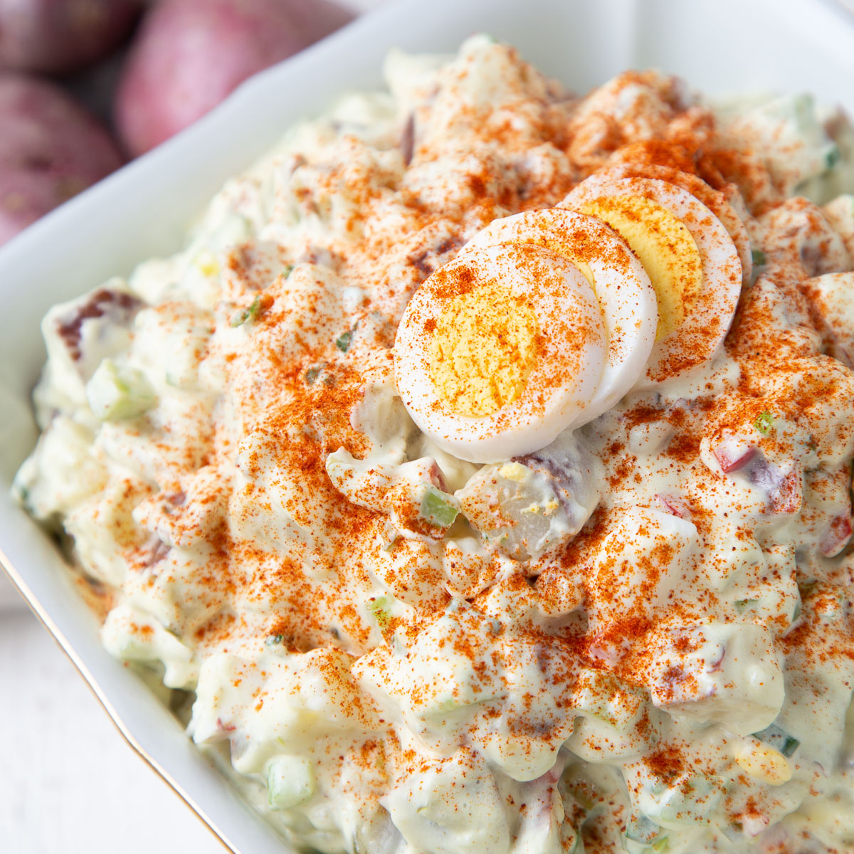 red potato salad topped with hard boiled eggs and paprika in a white dish.