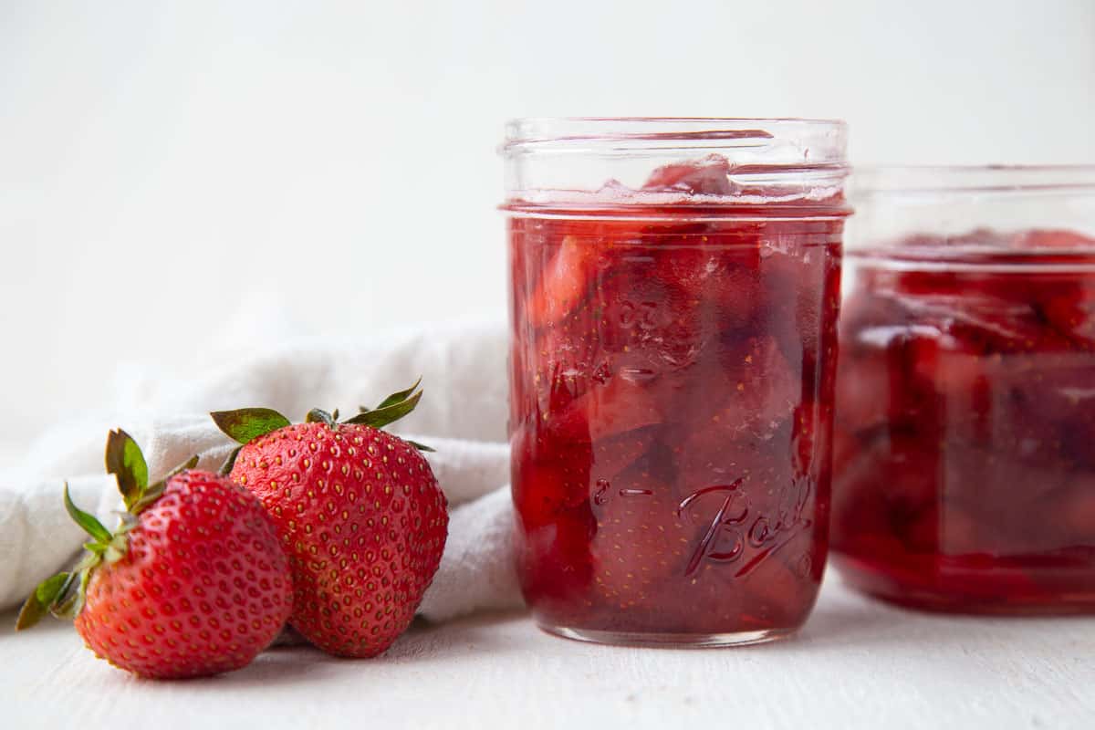 strawberry preserves in glass jars next to two fresh strawberries.