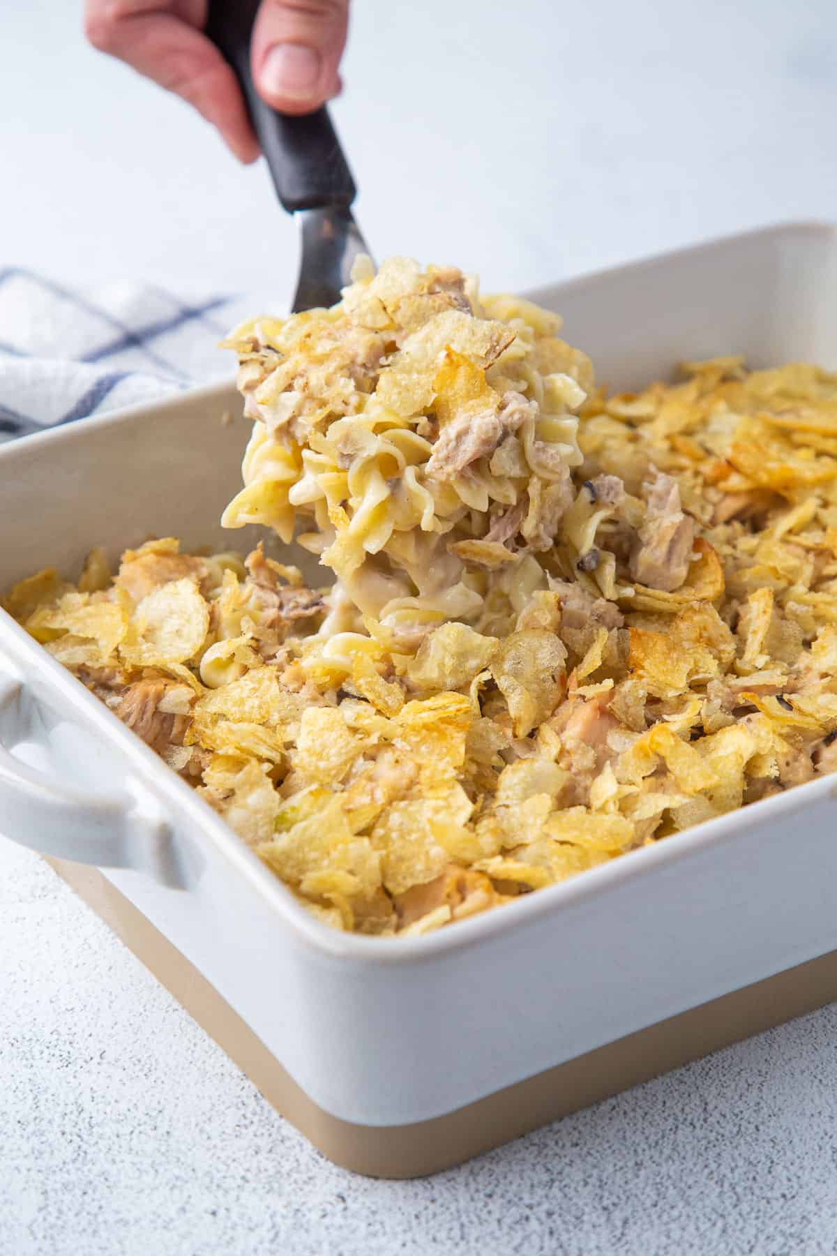 hand lifting a scoop of tuna noodle casserole out of a casserole dish.