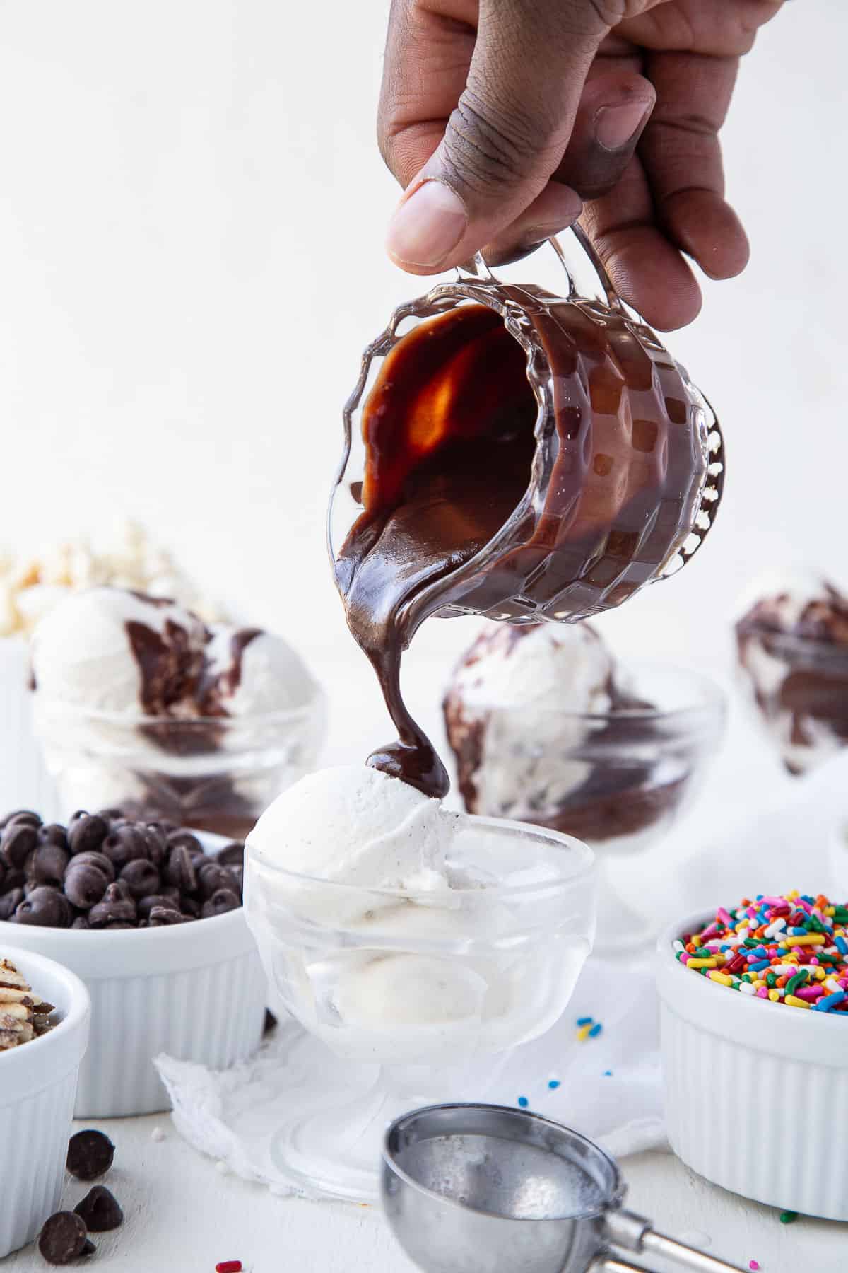 hand pouring hot fudge on a scoop of ice cream in a glass parfait dish.