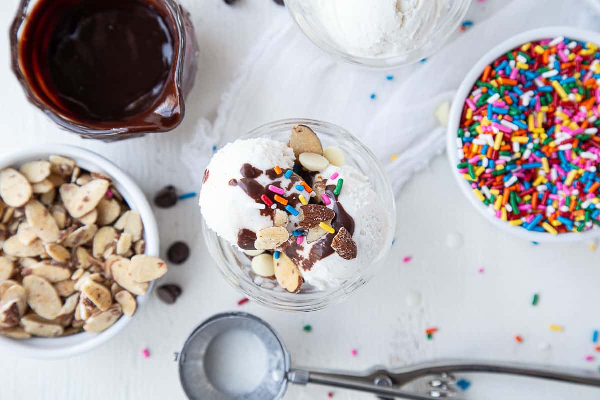 hot fudge sundae surrounded by bowls of fudge, sprinkles, and nuts.