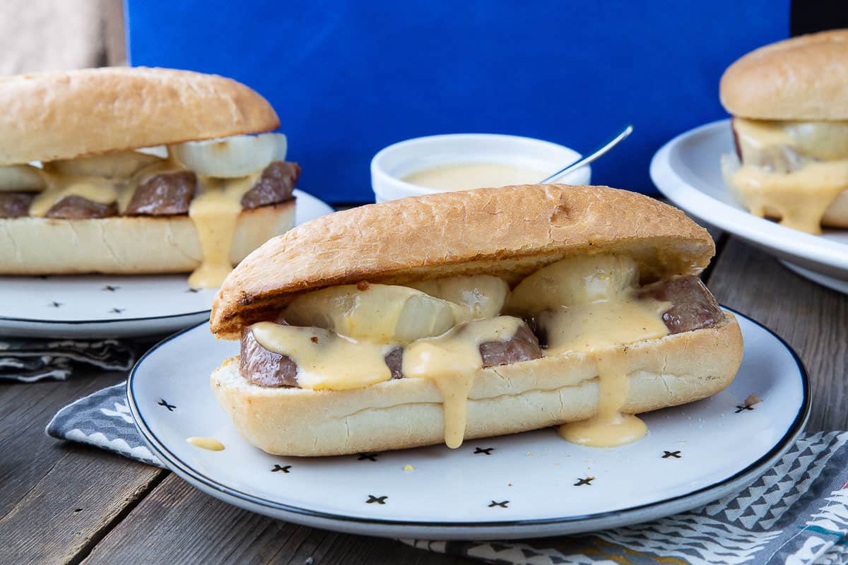 beer brat on a plate in a bun with onion slices and cheese sauce dripping out.