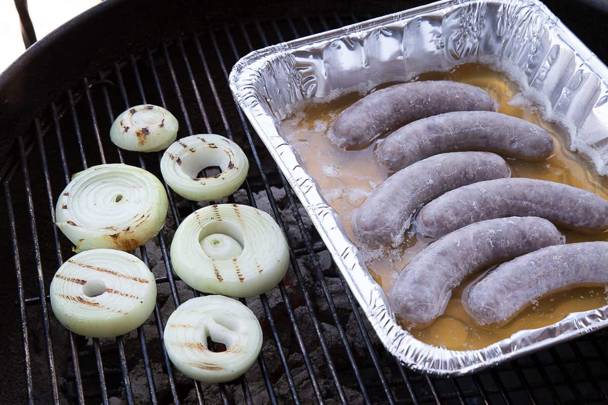 brats in a foil container with beer, plus sliced onions, on a grill.