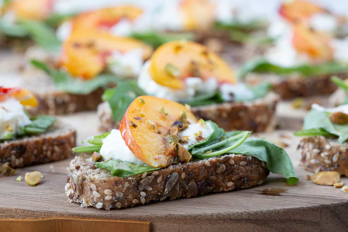 pieces of toast on a wooden board topped with arugula, burrata, peaches, and pistachios.