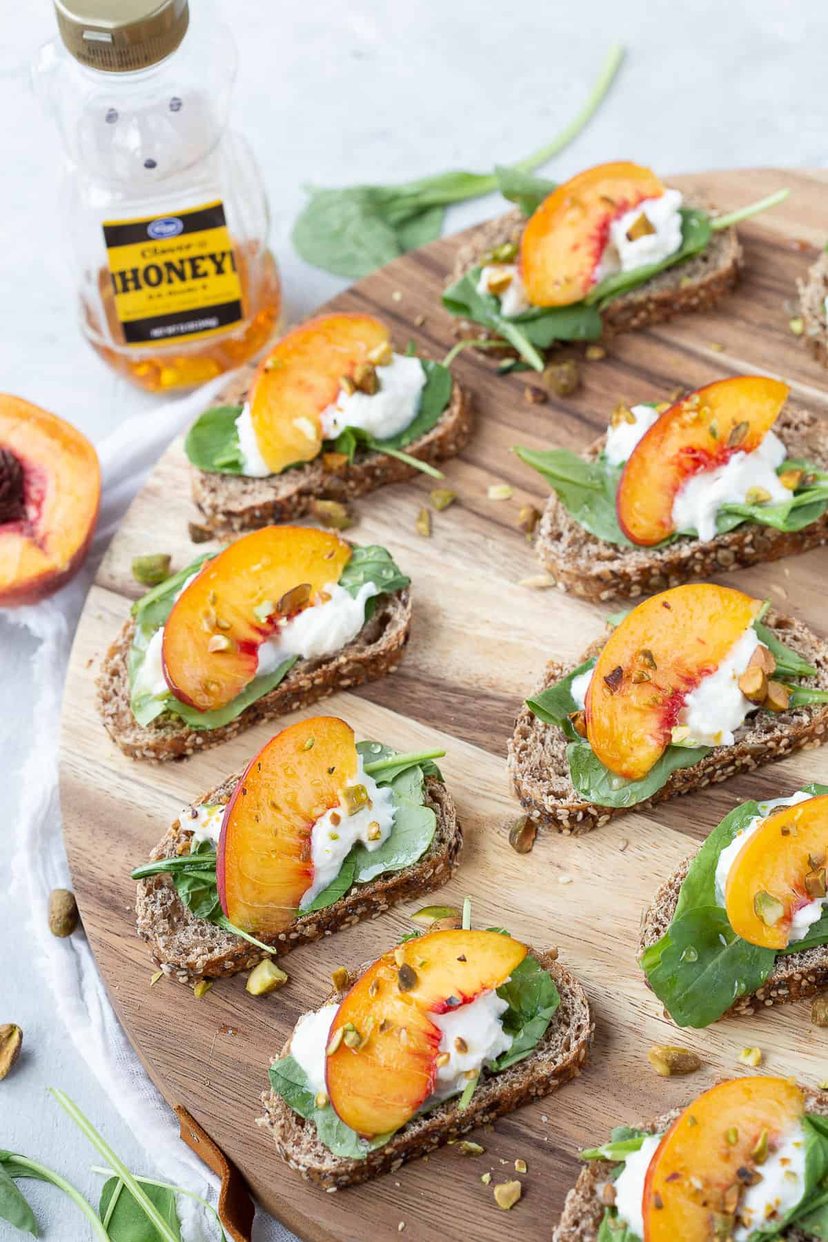 a jar of honey next to a wooden board of toasts topped with burrata, arugula, peaches, and pistachios.