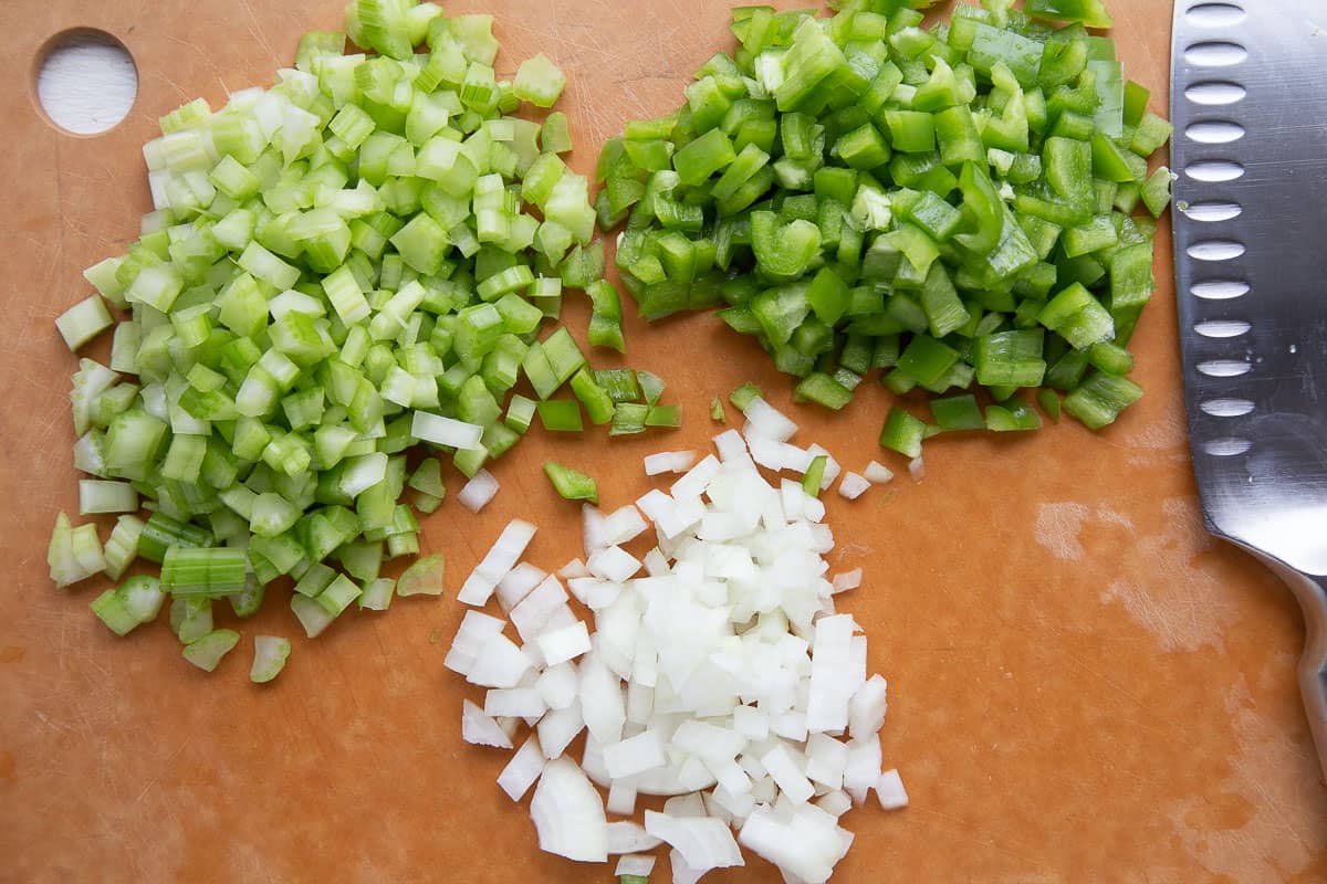chopped onion, celery, and green pepper on a brown cutting board next to a knife.