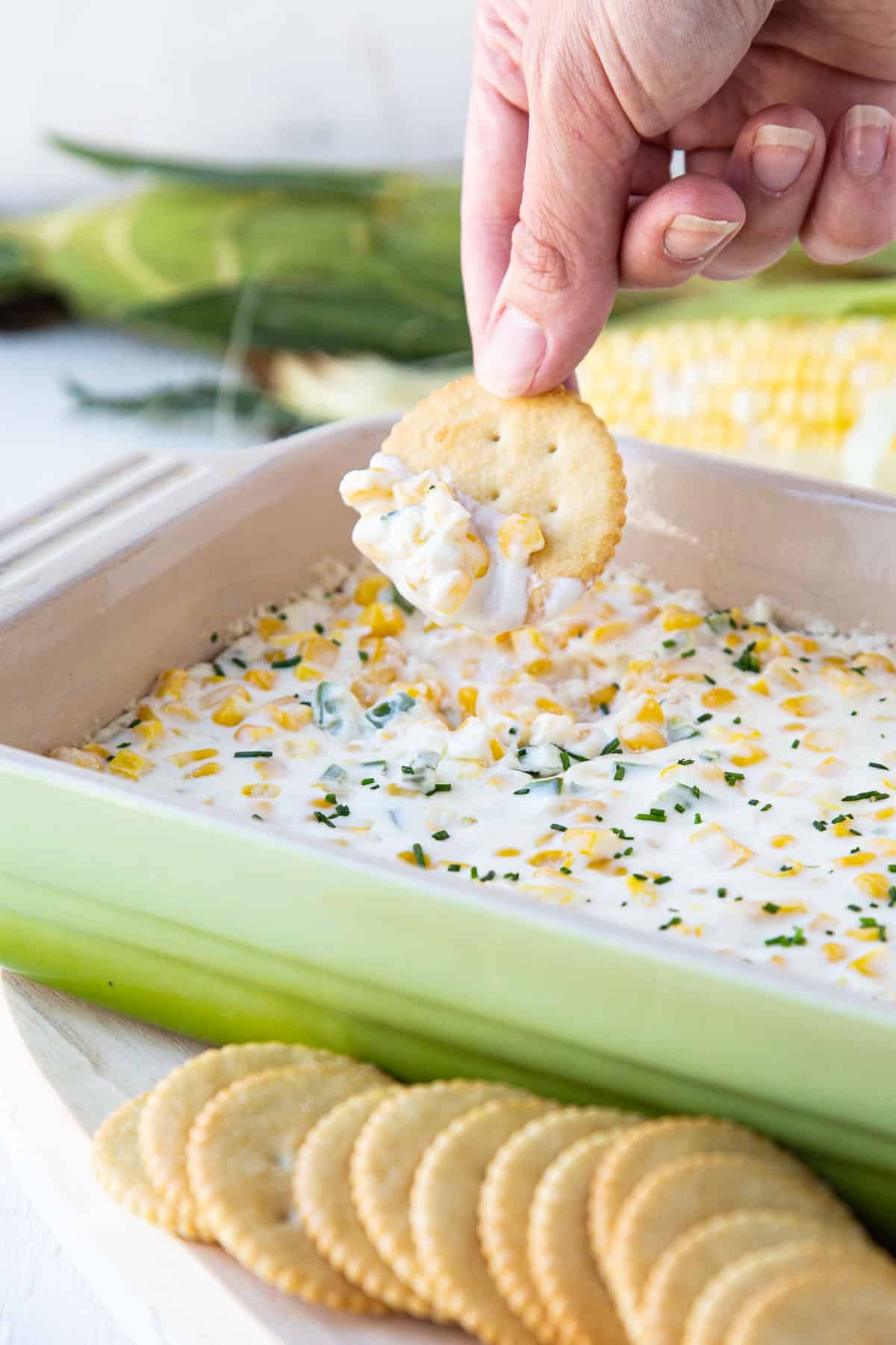 hand dipping a cracker into cream cheese corn dip in a green baking dish.