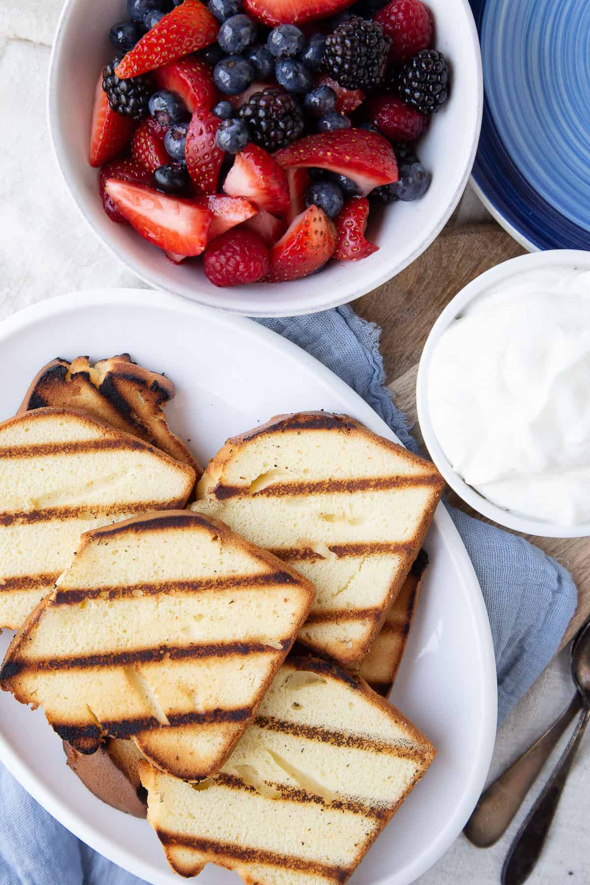 slices of grilled pound cake on a white platter, next to bowls of fresh berries and whipped cream.