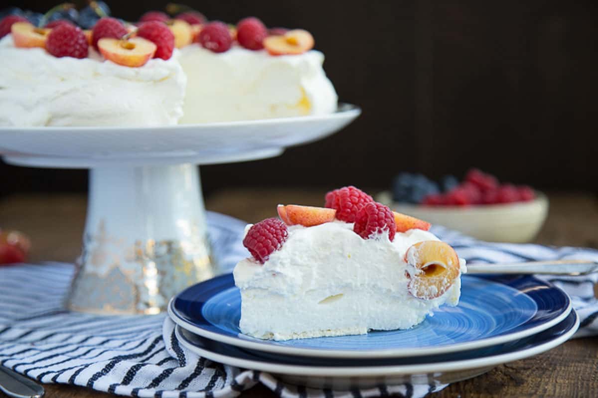 slice of berry pavlova on a blue plate next to a cake stand with a whole pavlova.