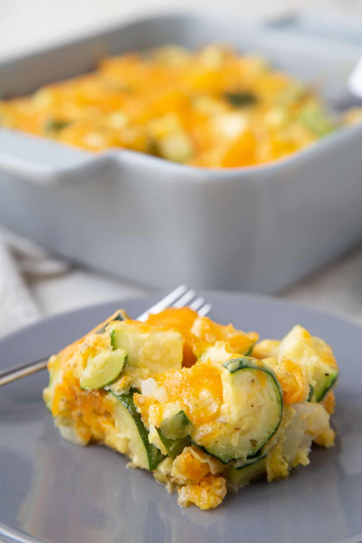 square slice of cheesy zucchini casserole on a gray plate with a gray casserole dish with more casserole in the background.