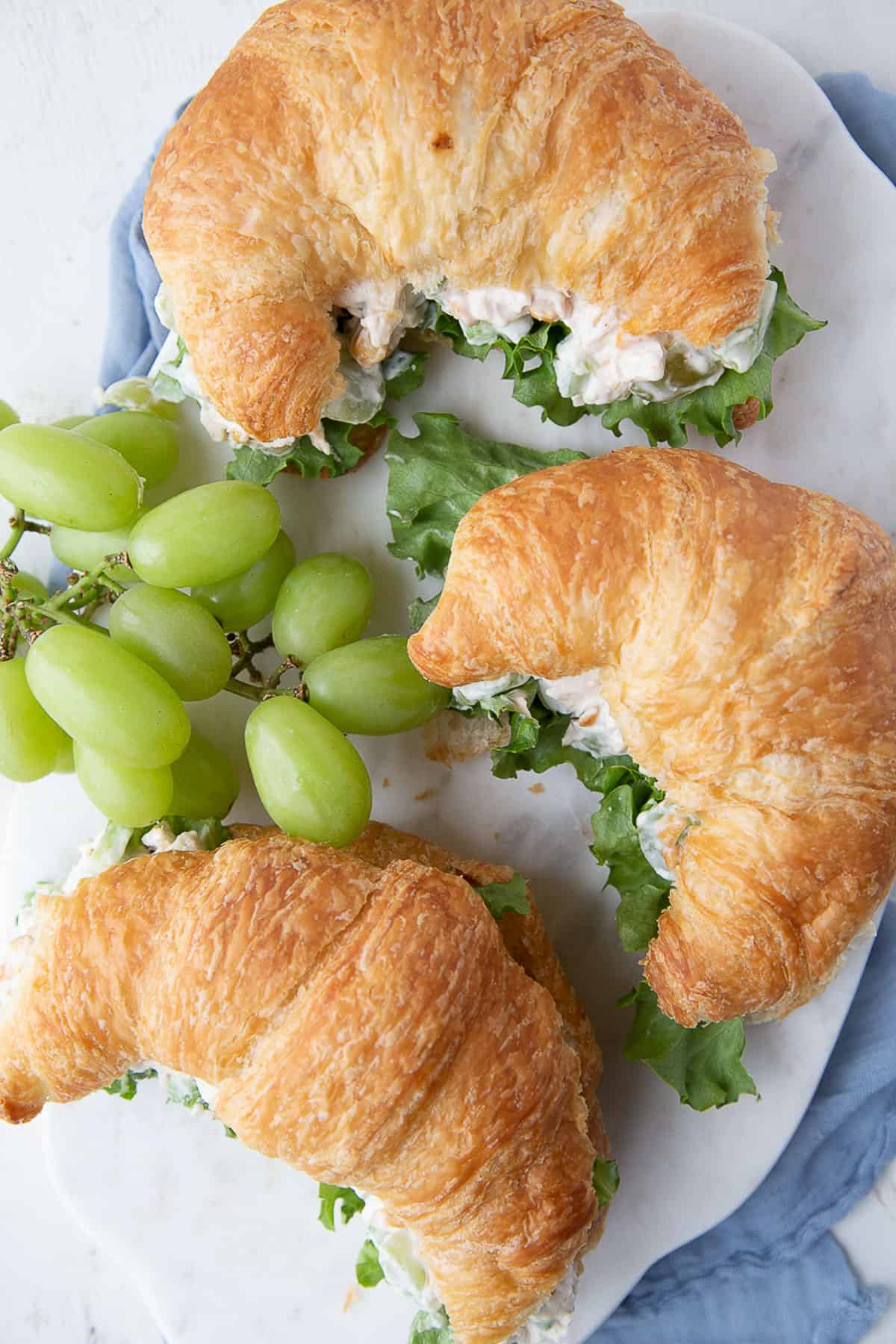 three croissants filled with chicken salad, next to a bunch of green grapes on a white platter.