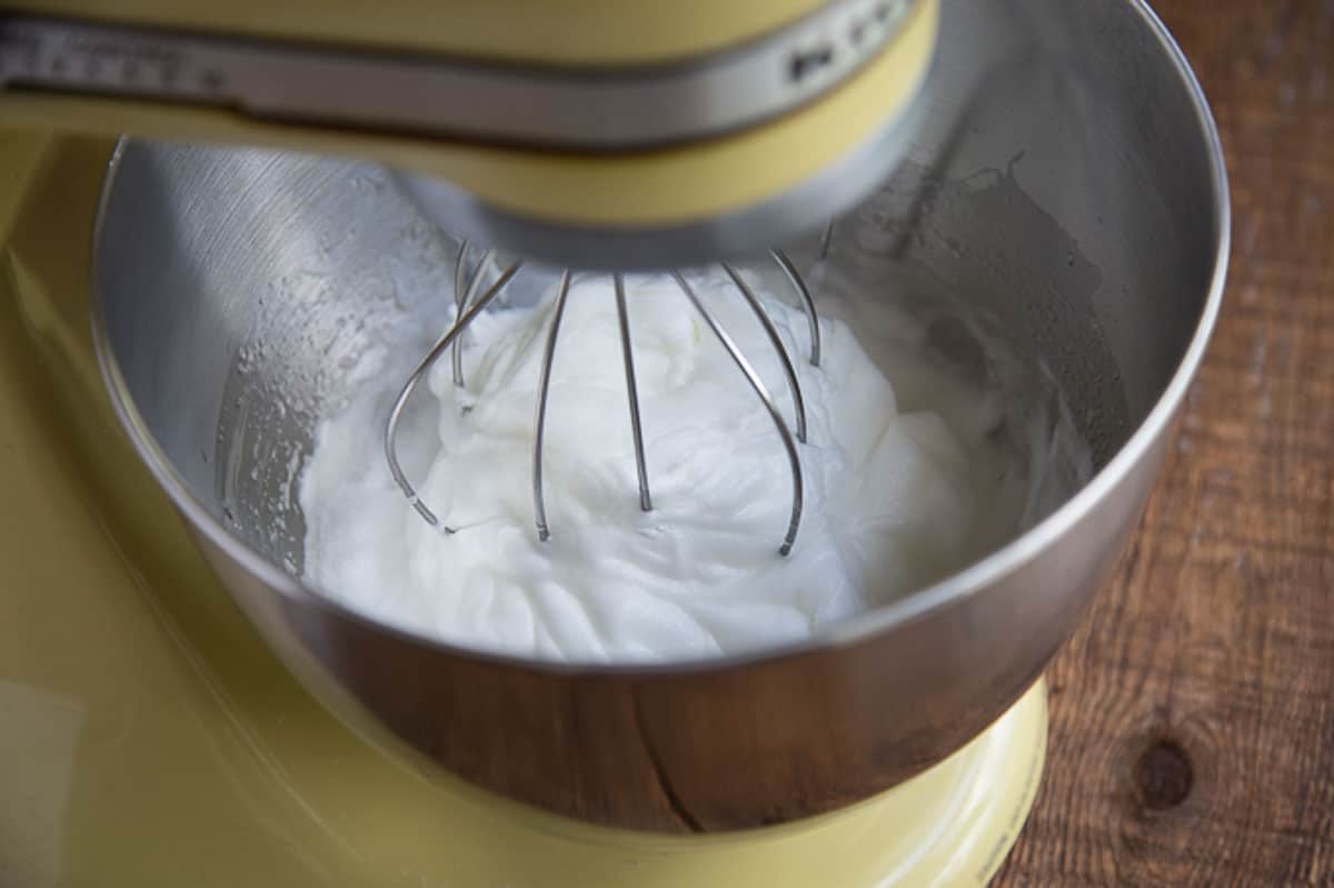 whipped egg whites in a metal bowl of a yellow stand mixer.