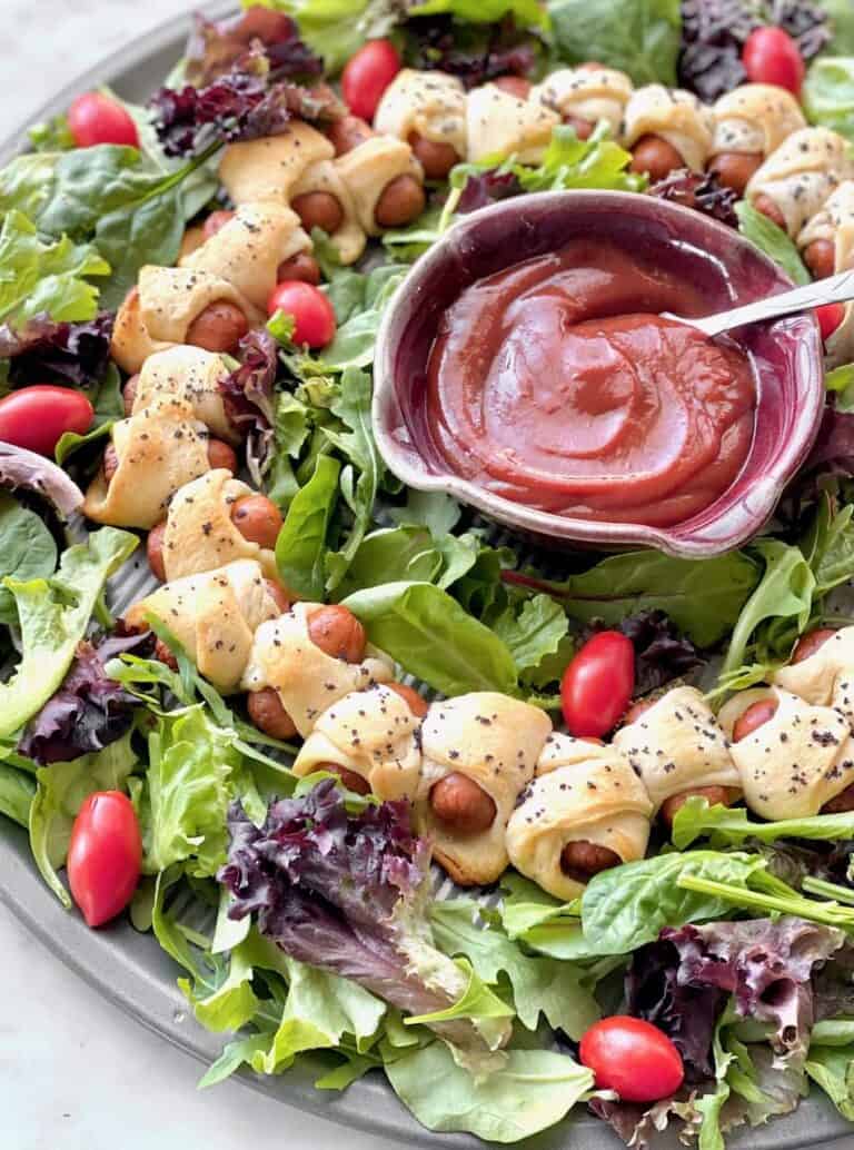 pigs in a blanket arranged in a circle on a bed of greens to resemble a wreath.