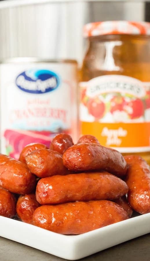 little smokies on a platter next to a jar of apple jelly and a can of cranberry sauce.