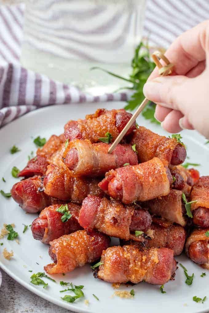 bacon wrapped sausages on a white platter, next to a hand piercing one with a toothpick.