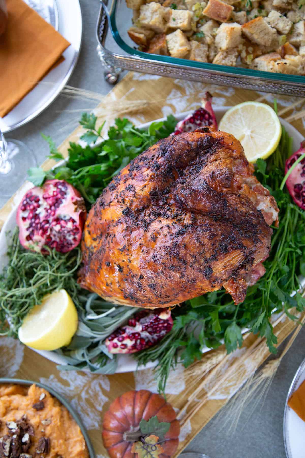 bone-in turkey breast on a platter of herbs with lemon halves, sitting on a table with stuffing and mashed sweet potatoes.