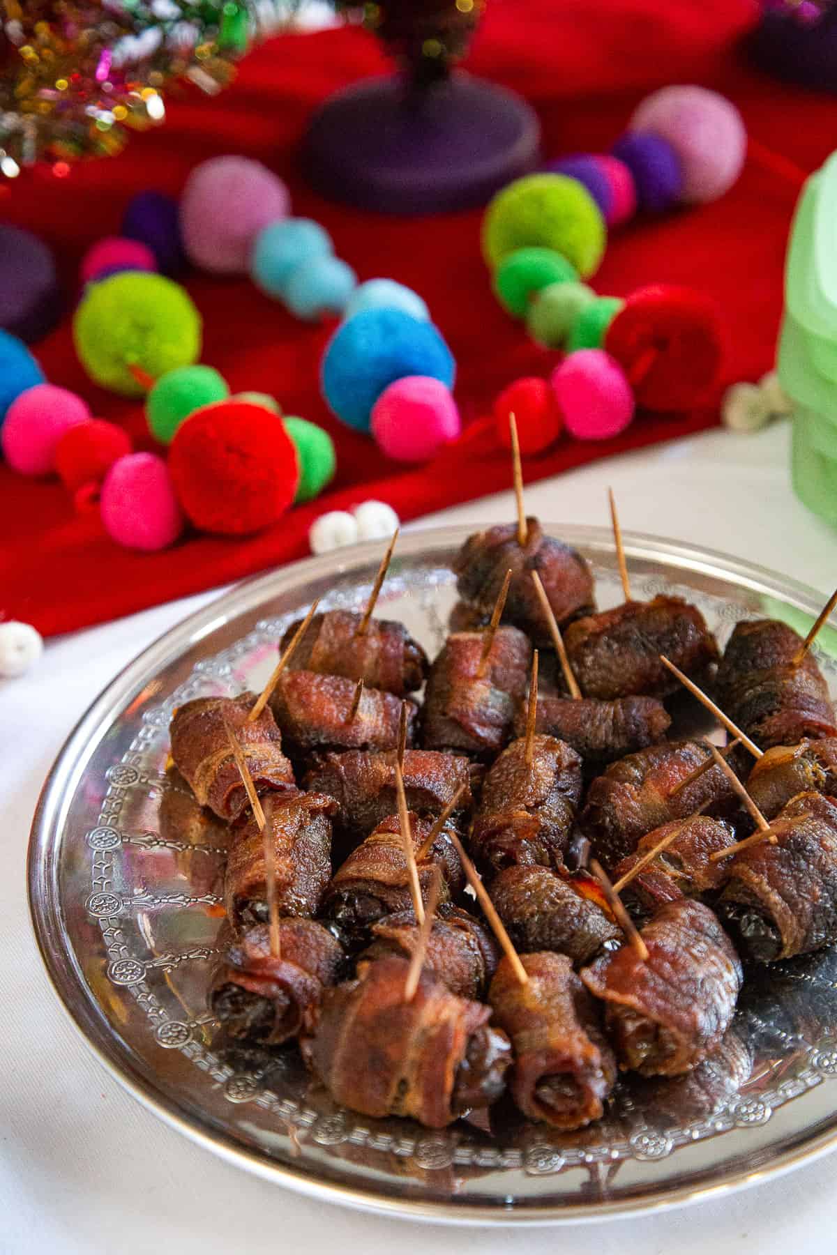 boozy bacon wrapped dates secured with toothpicks on a metal plate, next to colorful pompom garland.