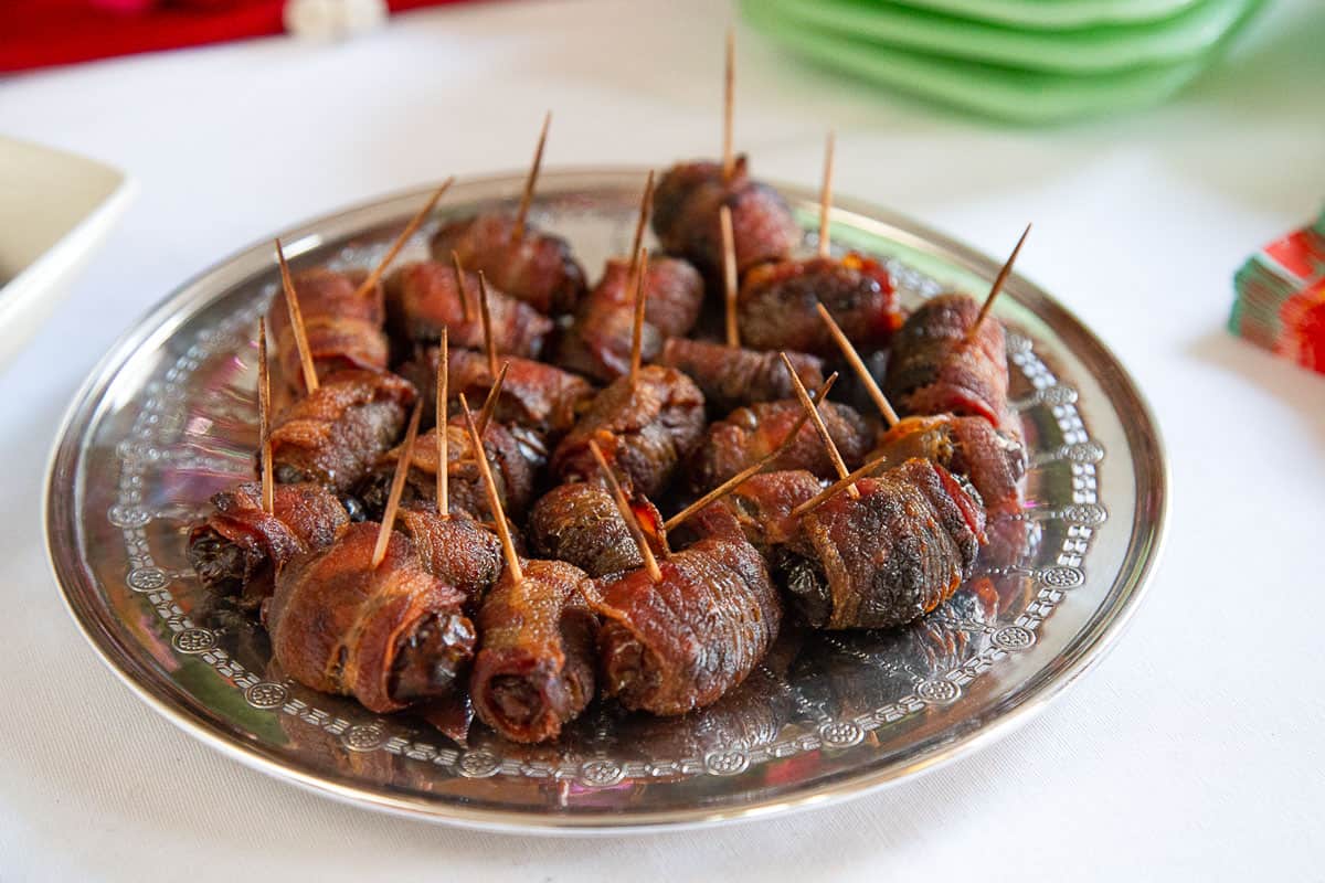 bacon wrapped dates on a round metal plate on a white tablecloth.