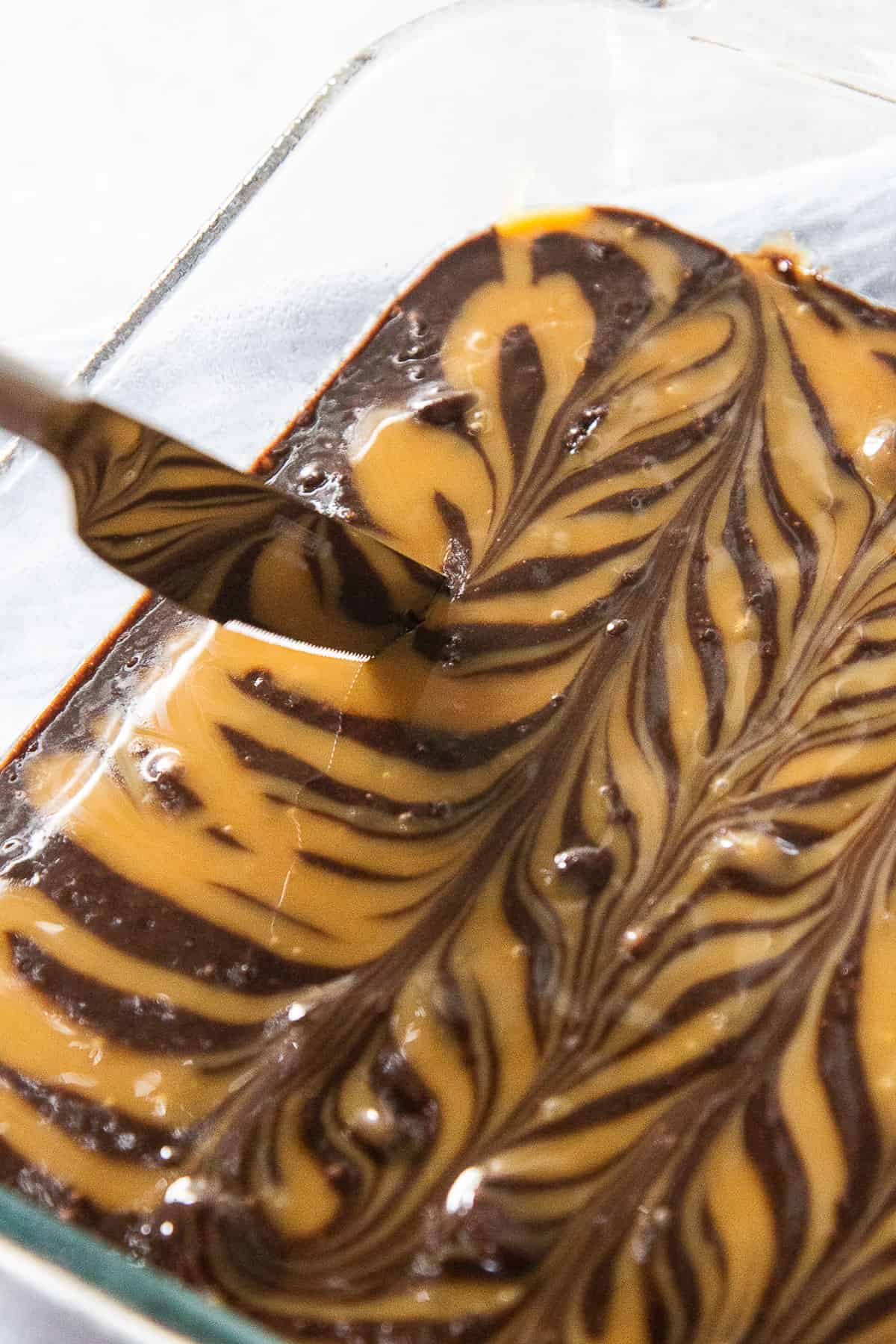a knife swirling caramel sauce into brownie batter in a glass pan.