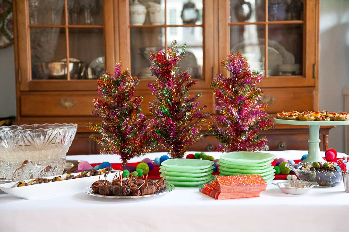 table with assorted appetizers, green plates, a punch bowl, and colorful tabletop christmas trees.