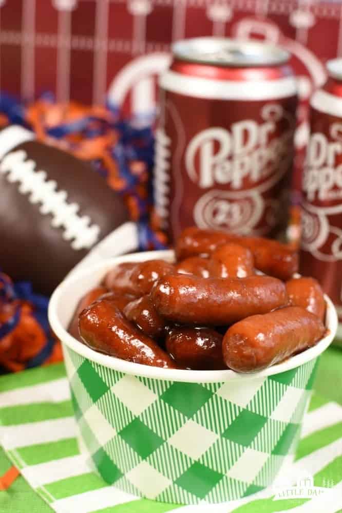 little smokies in a white and green cup next to a mini football and cans of dr pepper.