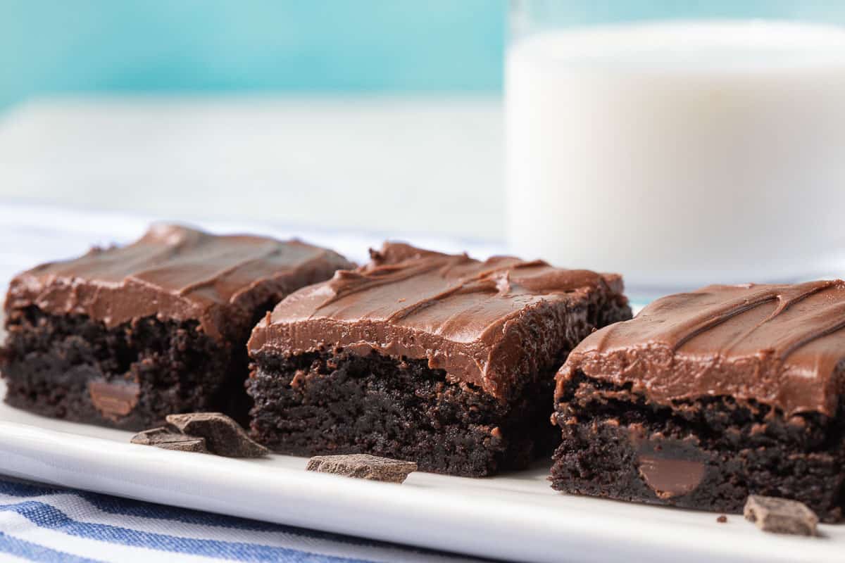 three brownies topped with chocolate frosting next to a glass of milk.