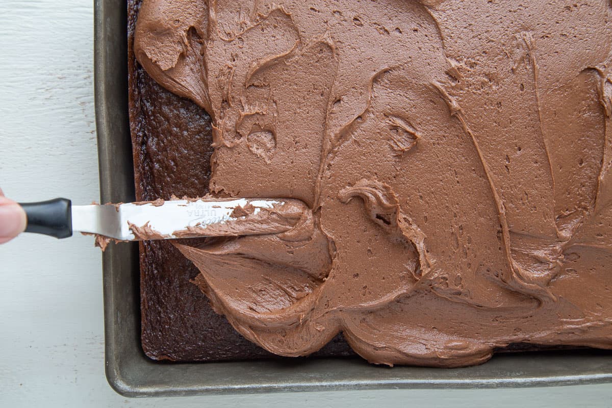 an offset spatula spreading chocolate frosting onto a chocolate cake.