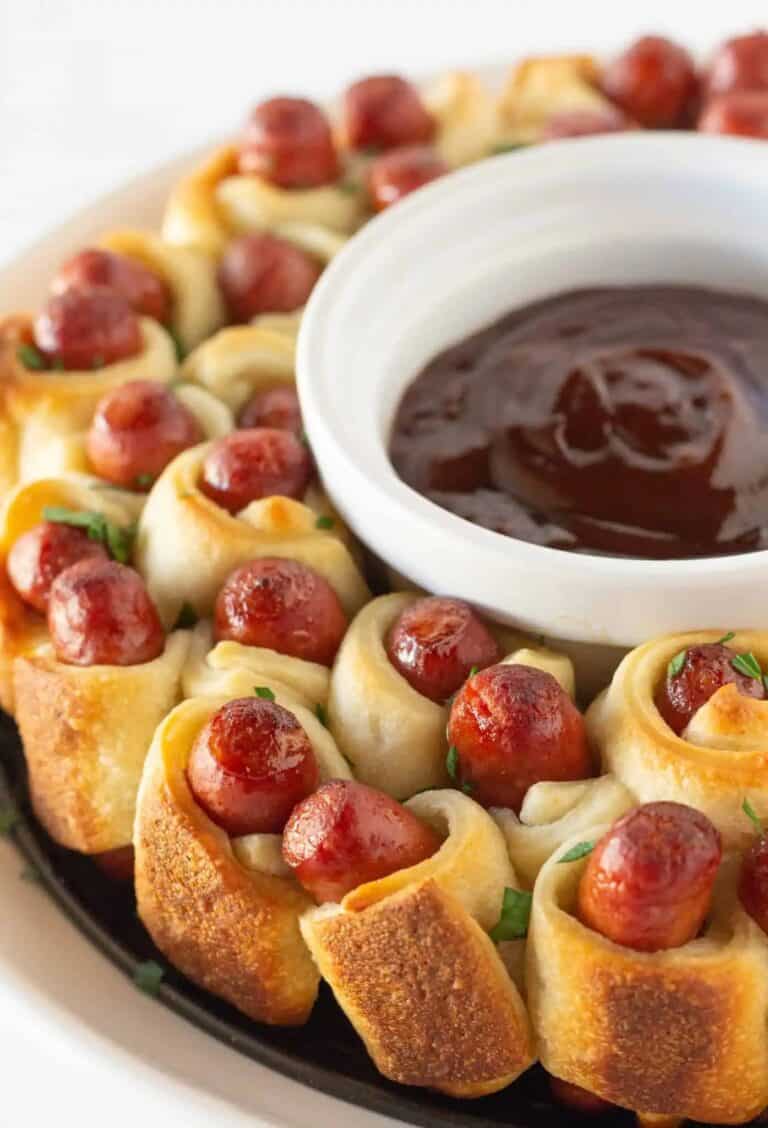 pigs in a blanket standing on their ends, arranged to resemble a wreath around a bowl of bbq sauce.