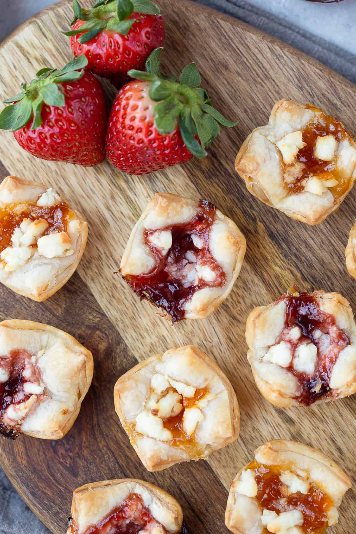 bite size puff pastry appetizers with goat cheese and jam inside on a wooden cutting board.