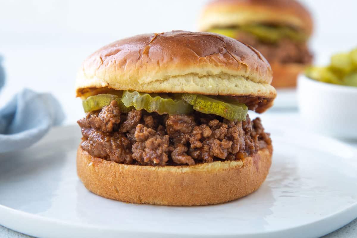 old fashioned sloppy joe sandwich on a brioche bun with pickles on a white plate.