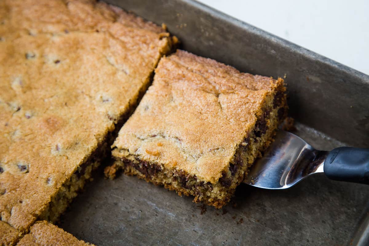 spatula scooping a chocolate chip cookie bar out of a metal pan.