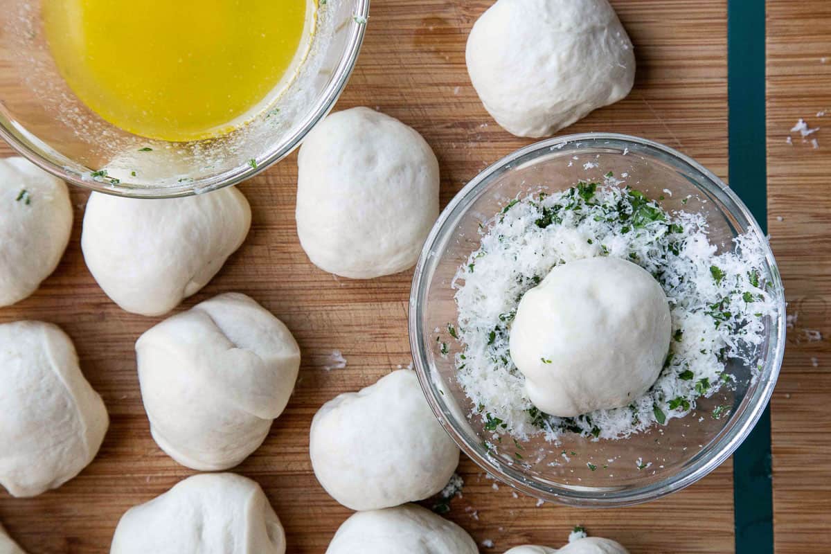dough balls on a wooden cutting board along with a shallow bowl of butter and a shallow bowl filled with parmesan and parsley.