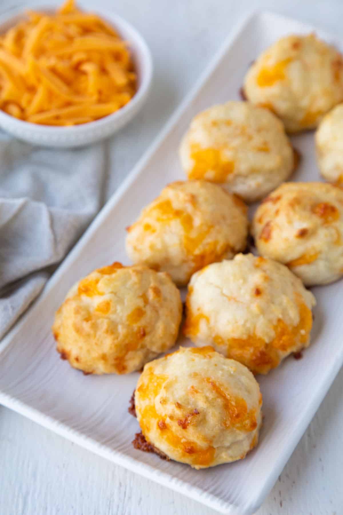 cheddar biscuits on a white rectangular serving platter next to a small bowl of shredded cheddar.