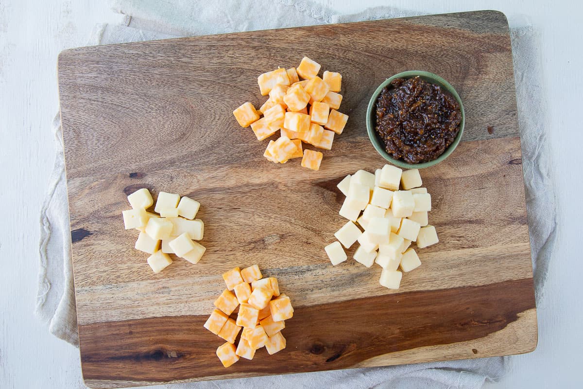 piles of cheese cubes and a small dish of fig jam on a wooden board.