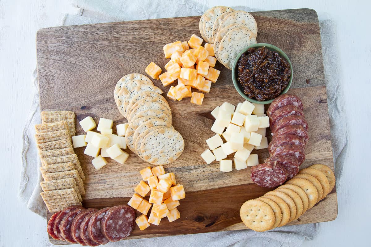 cheese cubes, crackers, salami, and a small dish of fig jam on a wooden board.