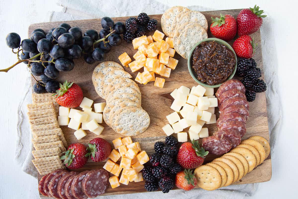 cheese cubes, salami, crackers, fruit, and a small dish of fig jam on a wooden board.