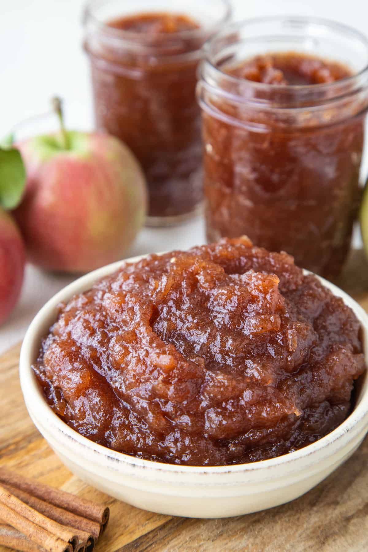 crockpot apple butter in a small yellow dish next to fresh apples and mason jars filled with apple butter.