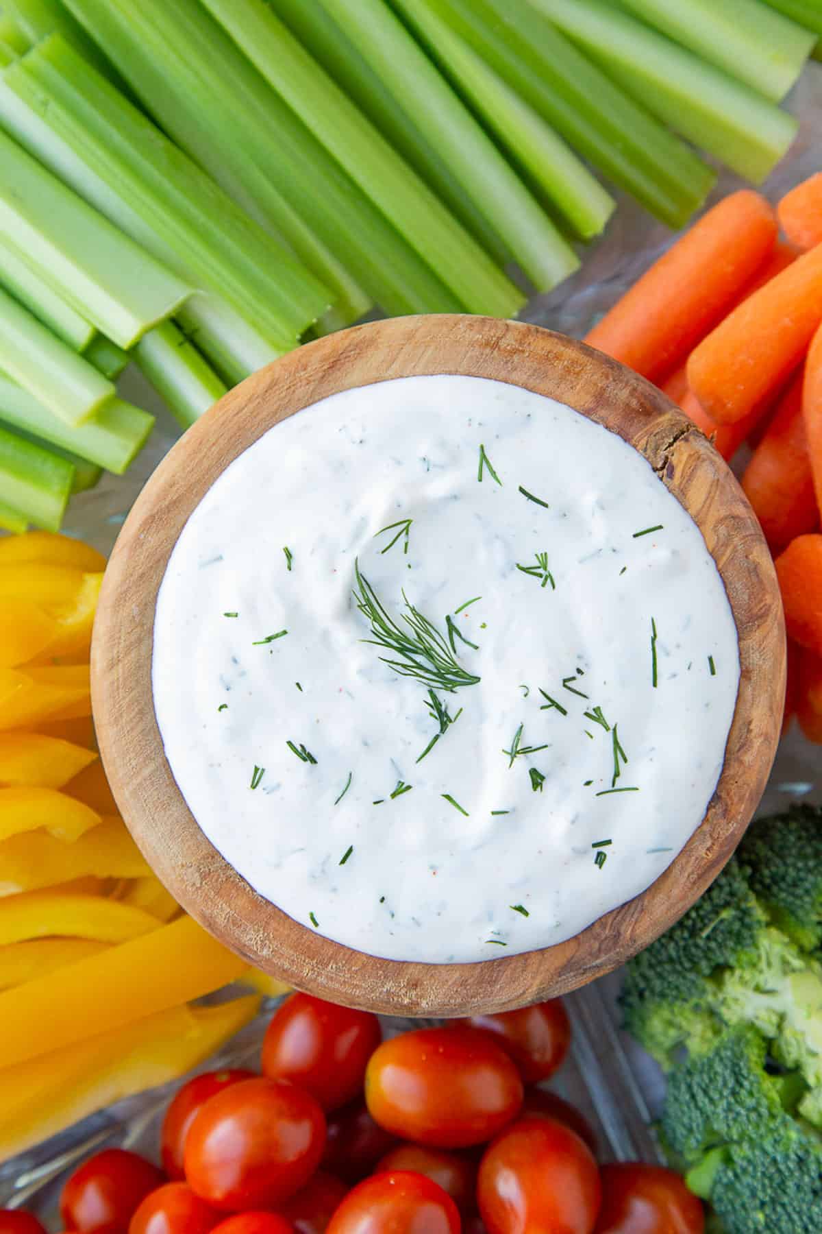white dill dip in a wooden bowl surrounded by assorted fresh vegetables.