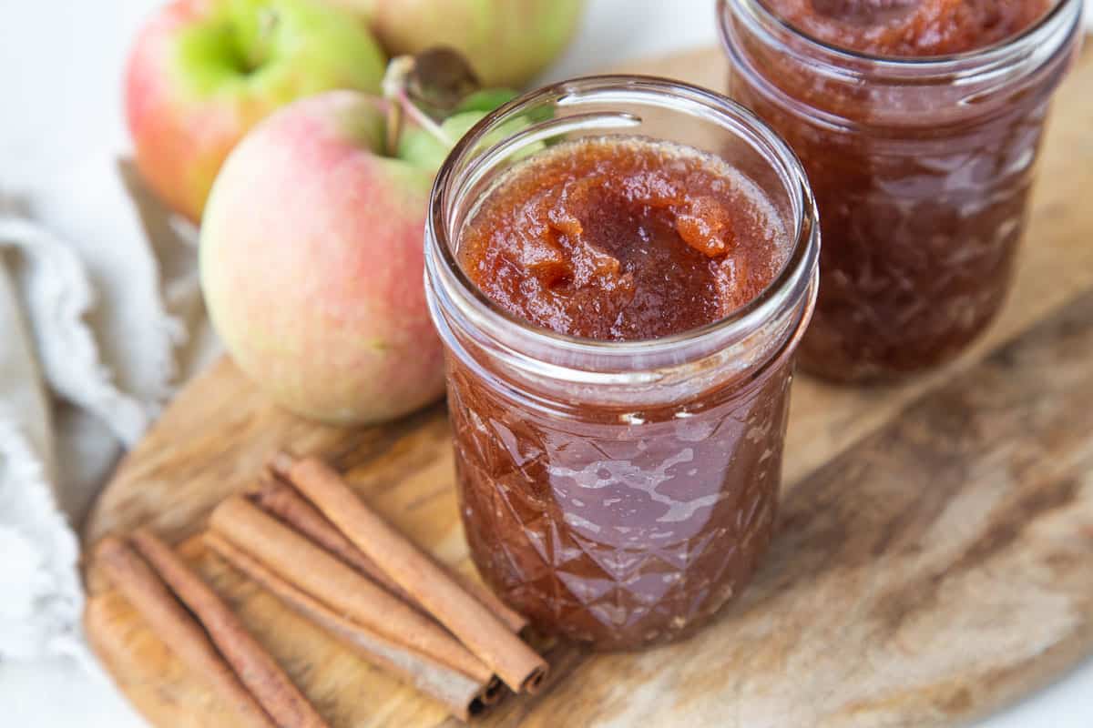 apple butter in mason jars next to cinnamon sticks and whole apples.