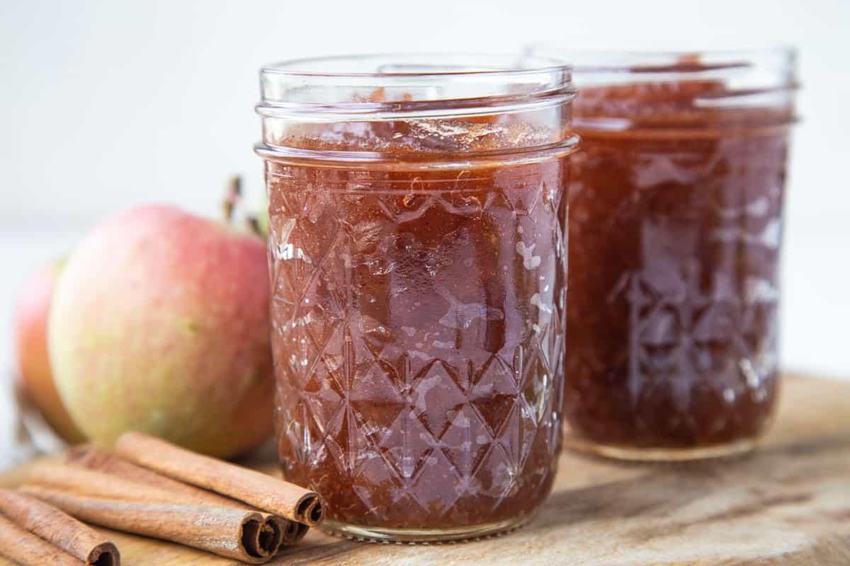 two mason jars filled with apple butter next to cinnamon sticks and a whole fresh apple.