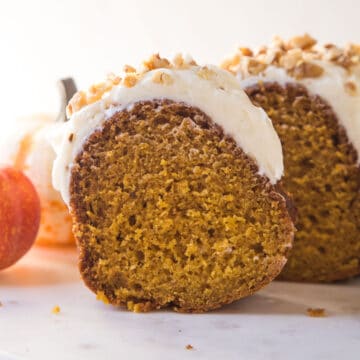 slice of pumpkin bundt cake with cream cheese frosting and chopped walnuts on top on a white marble platter.