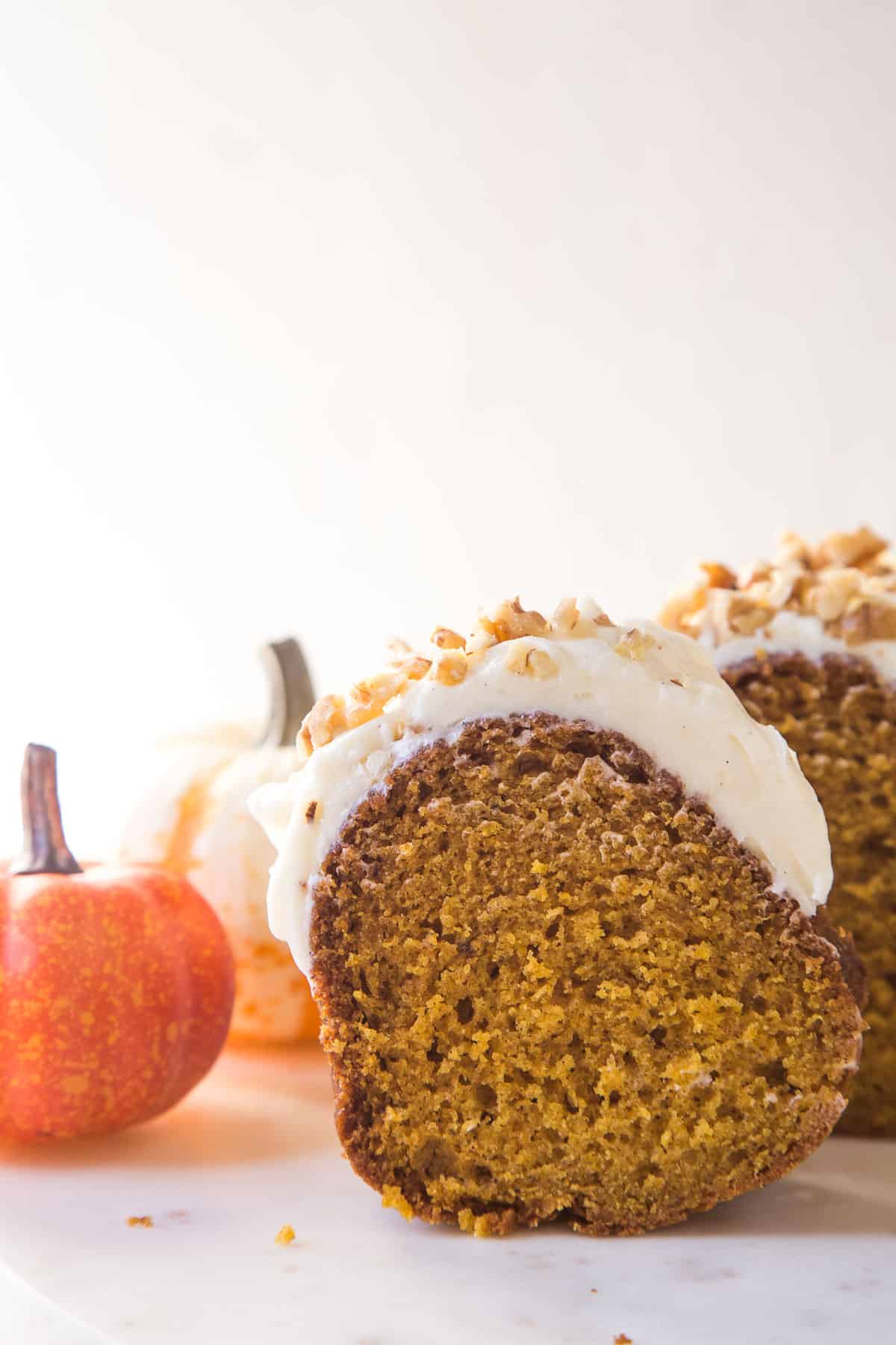 slice of pumpkin bundt cake topped with cream cheese frosting sitting next to two mini pumpkins.