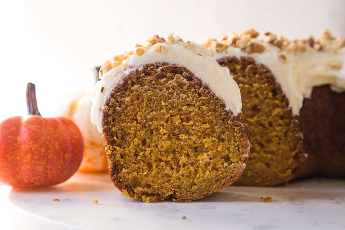 slice of pumpkin bundt cake topped with frosting and nuts, next to a mini pumpkin.
