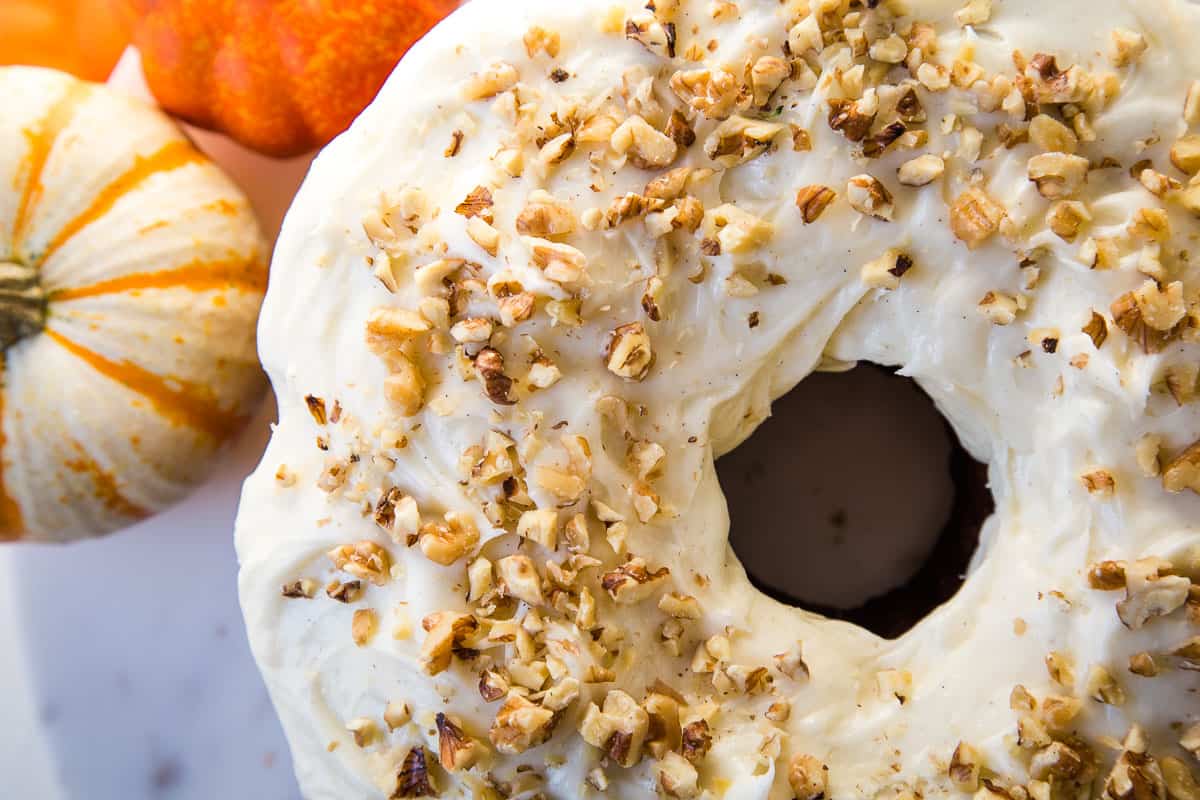 the top of a pumpkin bundt cake with cream cheese frosting and chopped walnuts next to two mini pumpkins.