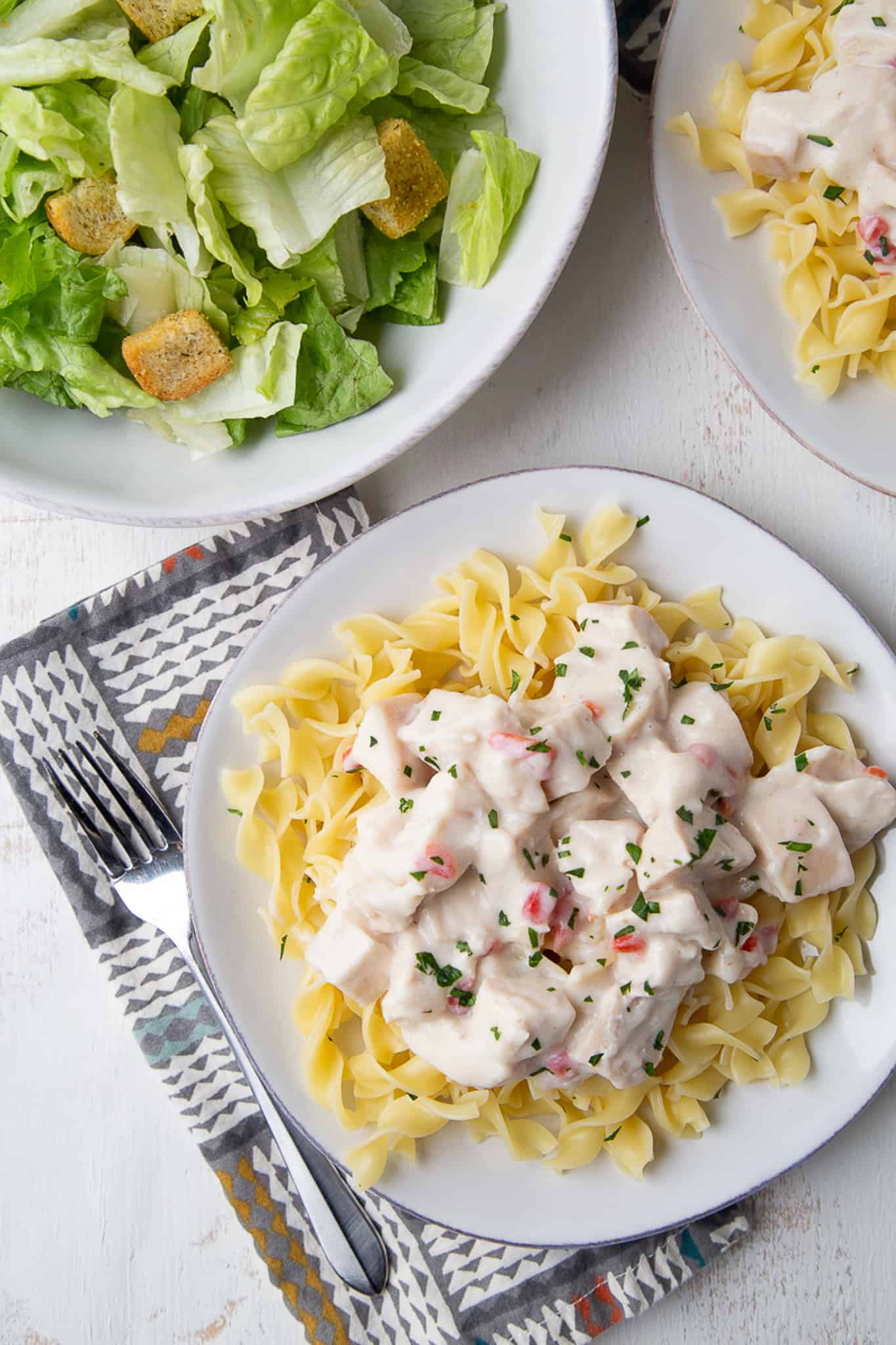 turkey in a cream sauce over egg noodles on a white plate, next to a plate of caesar salad.