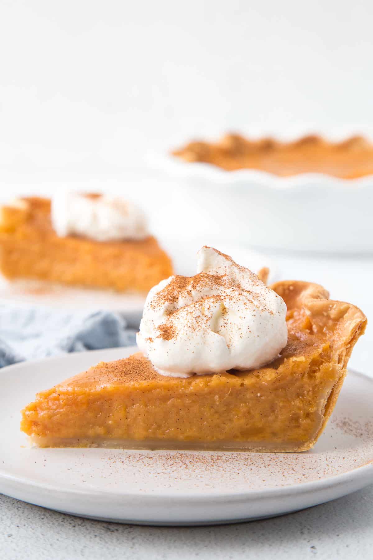 slice of old fashioned sweet potato pie on a white plate, topped with whipped cream and a sprinkle of cinnamon.