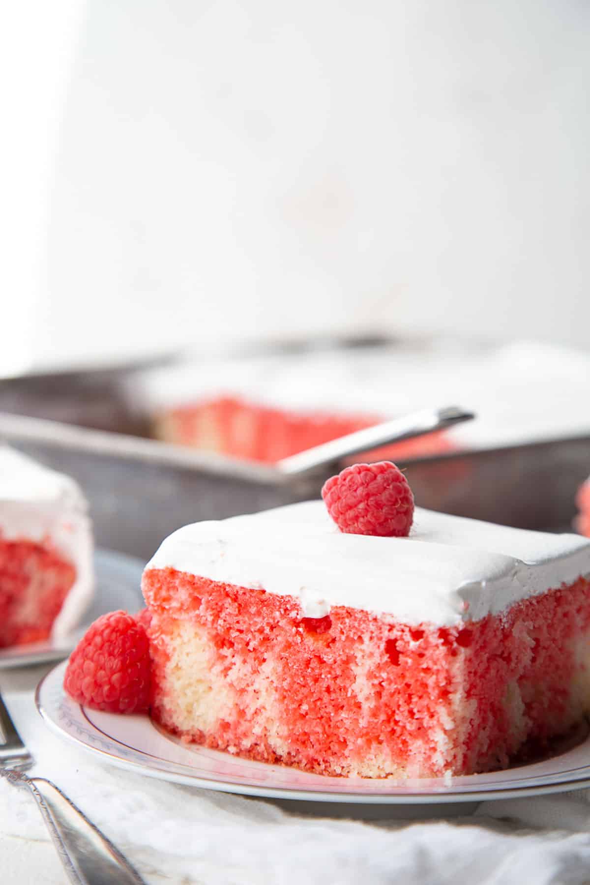 slice of jello poke cake with red jello, topped with a layer of whipped cream and a raspberry.