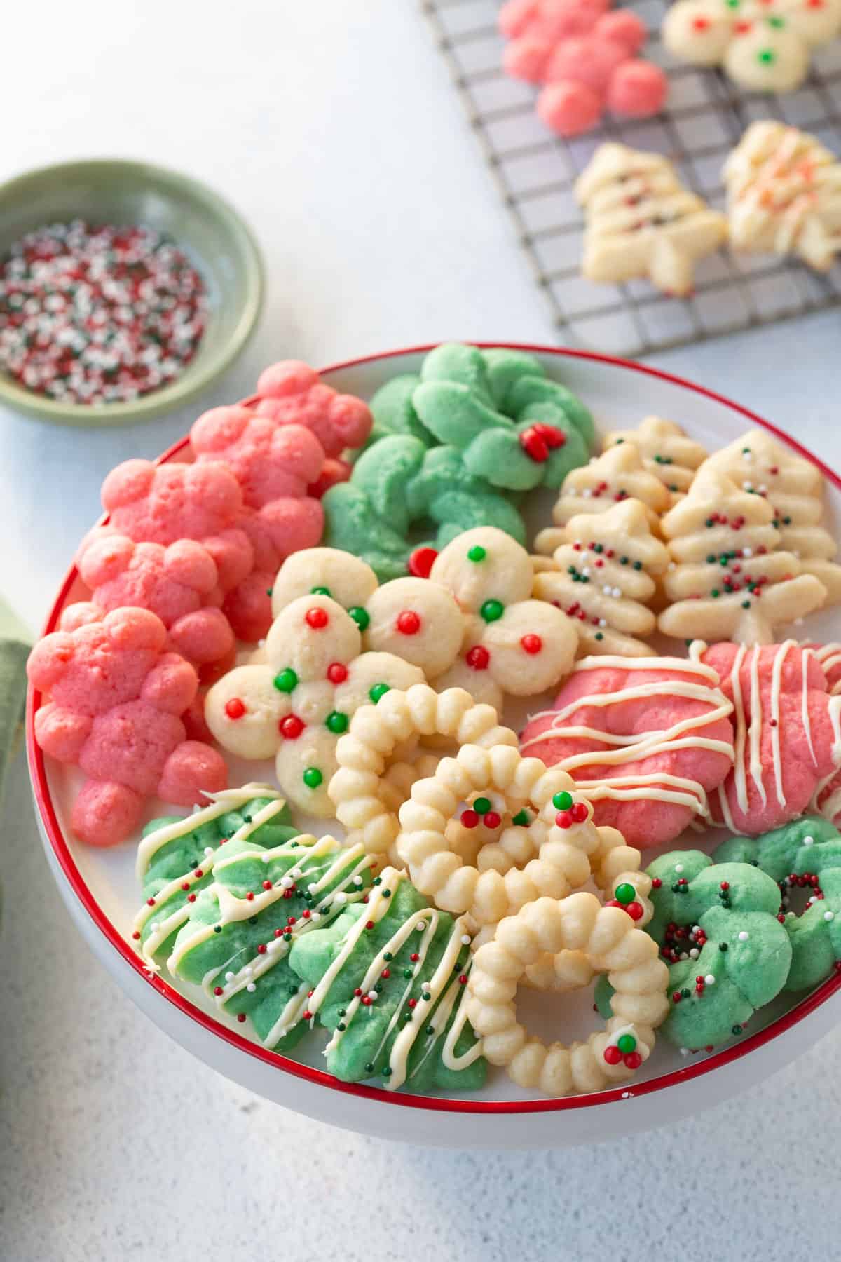 old fashioned spritz cookies on a white cake stand with a red rim, next to more spritz cookies on a wire rack.